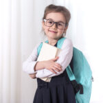 Girl-elementary school student with a backpack and a book with glasses is on a light background .The concept of education and primary school